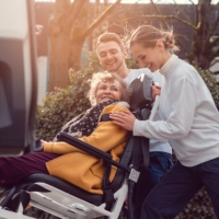 An elderly woman sitting on a wheelchair and being assisted by two persons