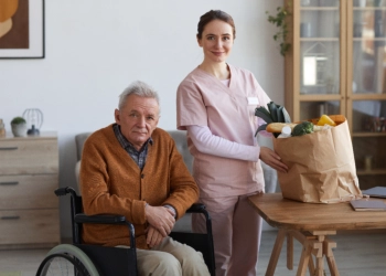 An elderly man sitting on a wheelchair and a female caregiver preparing a basket of fruits